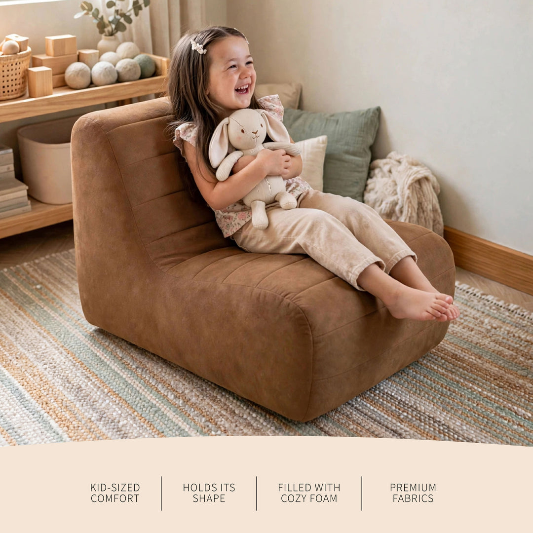 A young girl sits happily on a brown upholstered lounge chair in a styled nursery or playroom, holding a plush bunny toy. The scene includes a striped rug, wooden shelf, and decorative pillows in the background. 