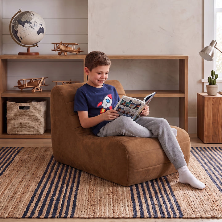 A young boy sits comfortably in a brown suede-like bean bag chair, reading a book in a cozy, well-lit room with a wooden bookshelf, globe, and striped rug. 