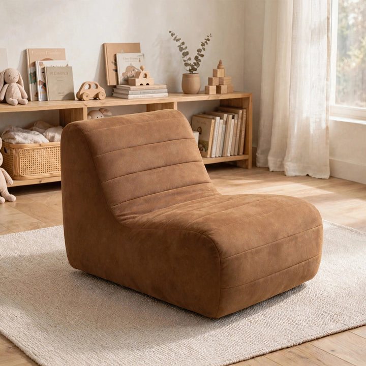 A plush, wedge-shaped lounge chair in a warm camel brown faux suede material, styled on a light beige rug in a bright, modern nursery or playroom setting. 