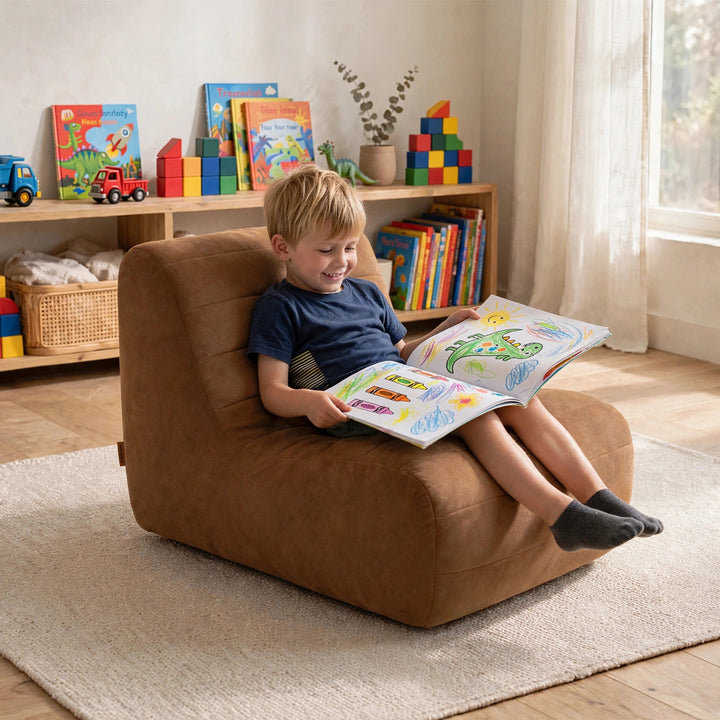 A young boy sits in a brown upholstered lounge chair reading a colorful book in a bright children&