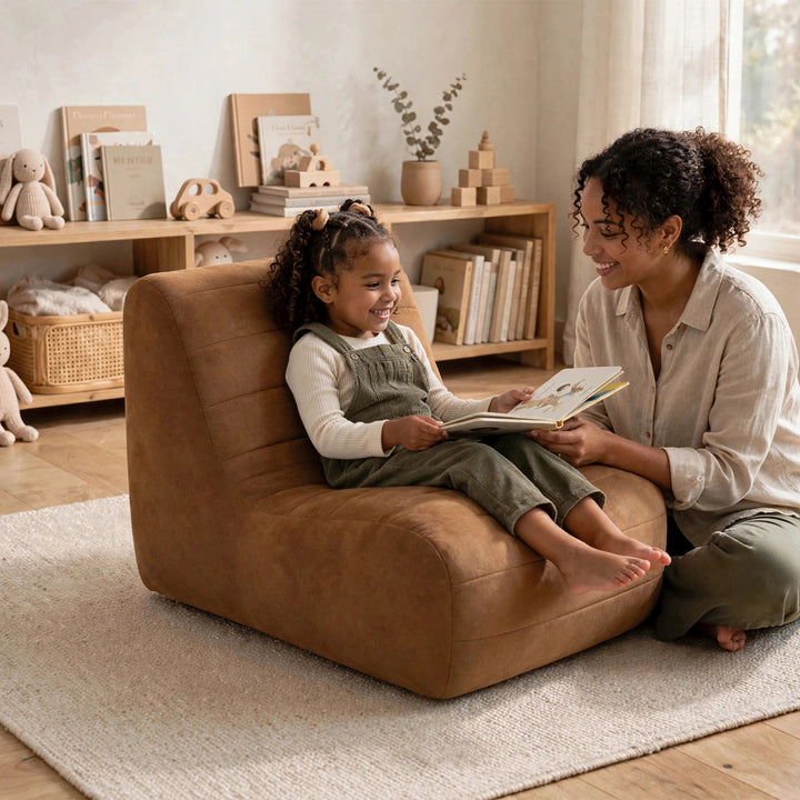 A woman and a young girl are reading a book in a cozy, warmly lit room. The girl is sitting in a plush, tan-brown upholstered lounge chair, while the woman kneels beside her on a light beige rug. 