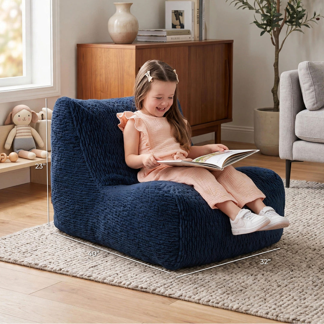 A young girl sits smiling on a plush navy blue lounge chair in a bright living room, reading a book. White dimension lines overlay the chair showing its measurements: 23 x 49 x 32 inches. 