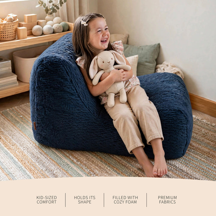 A young girl with long brown hair sits happily on a large, dark navy blue bean bag chair, holding a cream-colored stuffed bunny. She is wearing beige pajamas and barefoot on a striped woven rug in a cozy, sunlit room. 