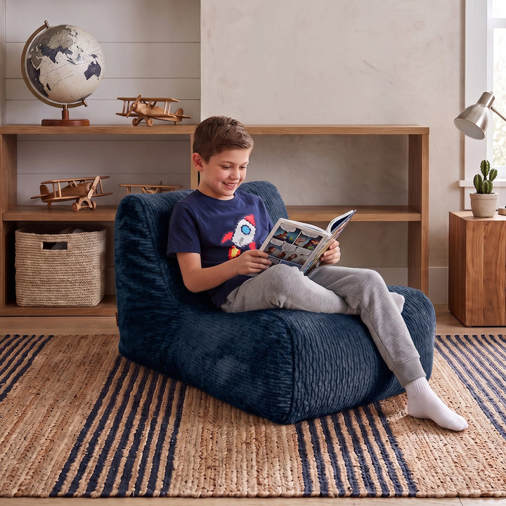 A child sitting on a navy blue bean bag chair in a cozy room setting, reading a book. The scene includes a wooden bookshelf with a globe and toy airplanes, a striped rug, and a side table with a lamp. 