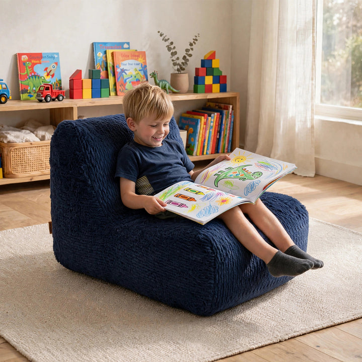 A young boy sits smiling in a dark navy blue textured bean bag chair, reading a colorful book in a bright children&