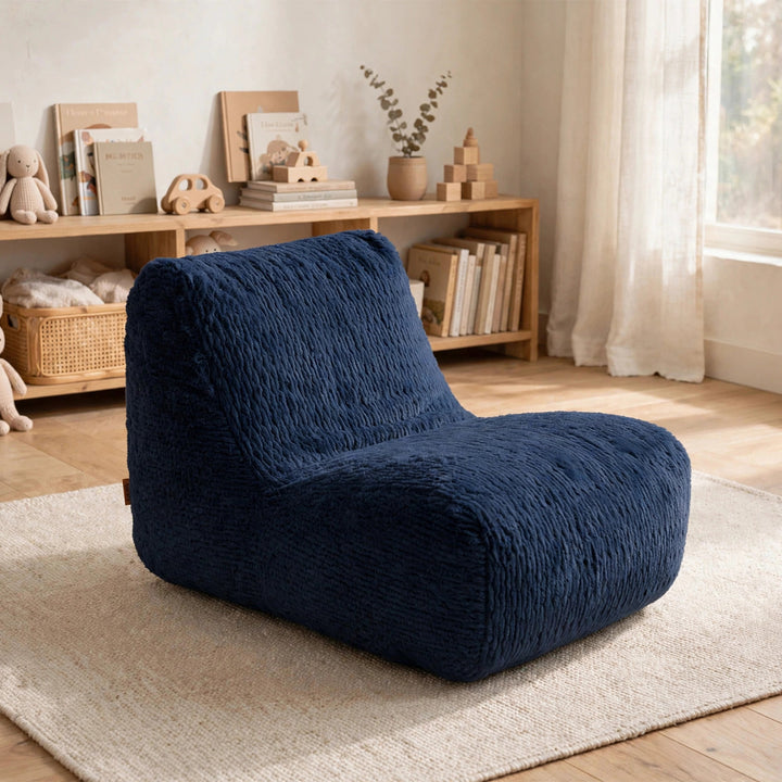 A navy blue textured fabric lounge chair placed on a light beige rug in a softly styled room with wooden shelves, books, toys, and a window with sheer curtains. 