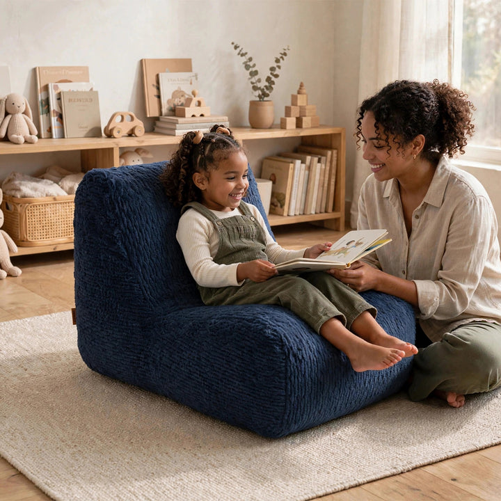 A young girl sits smiling in a textured navy blue floor chair while a woman reads a book to her in a bright, cozy room with wooden shelves and toys. 