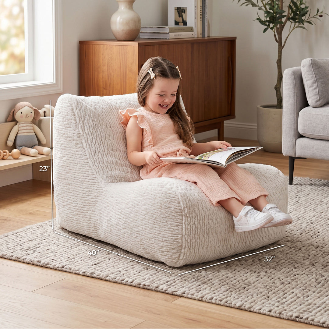 A young girl sits smiling in a cream textured armchair in a bright living room, reading a book. White dimension lines overlay the chair showing its 23 x 10 x 32 inch measurements. 