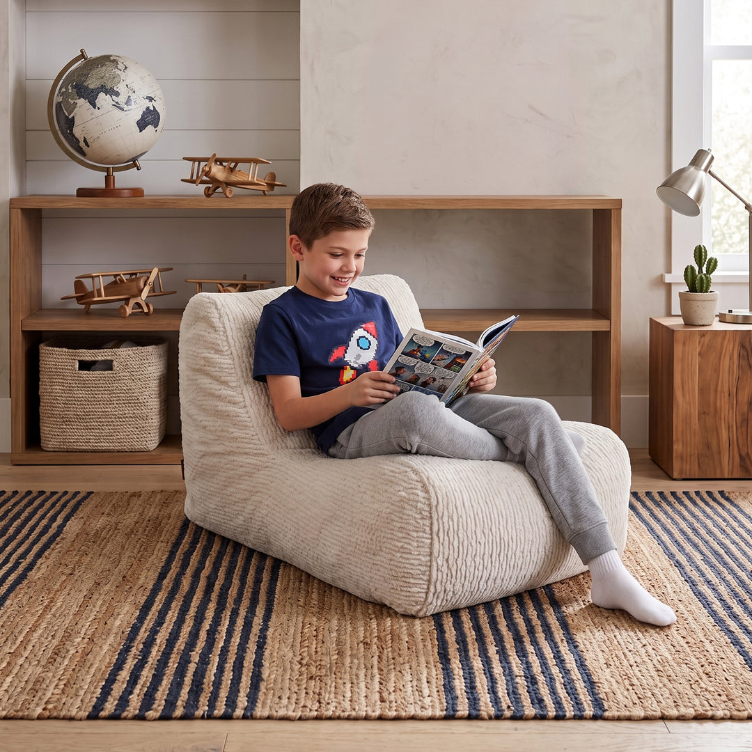A young boy sits comfortably in a cream-colored textured bean bag chair, reading a book in a cozy, well-lit room with wooden shelves, a globe, and a striped rug. 
