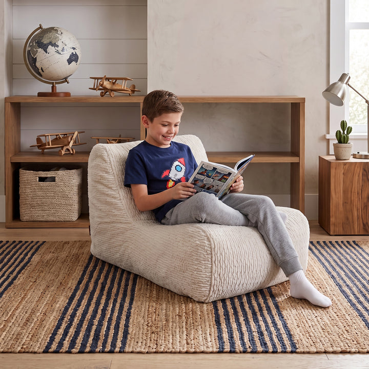 A young boy sits comfortably in a cream-colored textured bean bag chair, reading a book in a cozy, well-lit room with wooden shelves, a globe, and a striped rug. 