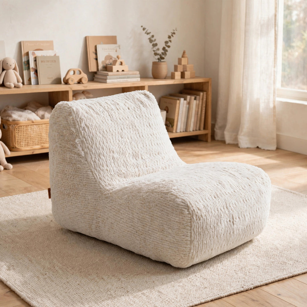 A plush, low-profile lounge chair upholstered in a textured cream bouclé fabric, set on a woven rug in a softly lit, neutral-toned room with a wooden bookshelf in the background. 