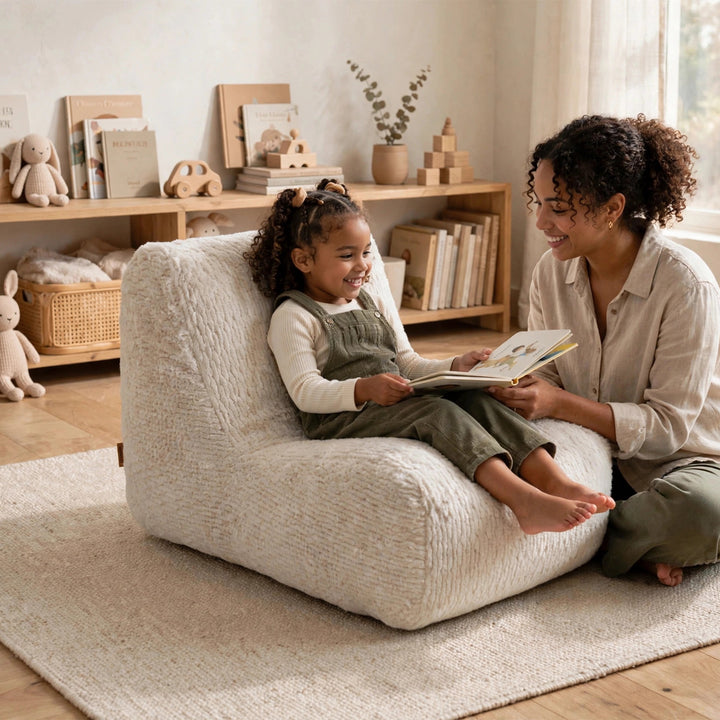 A woman and a young girl are reading a book together in a cozy, sunlit room. The girl is seated on a chunky, textured cream bouclé armchair, while the woman sits beside her on a light rug. 