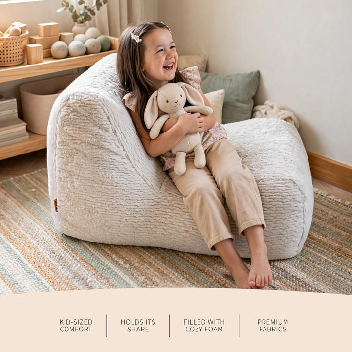 A young girl sits happily on a large, cream-colored textured bean bag chair in a cozy room, holding a plush bunny toy. The image features text overlays at the bottom highlighting the product&