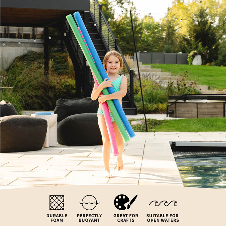 A young girl stands on a pool deck holding three long pool noodles in blue, green, and pink. The background shows a backyard patio, lounge chairs, a staircase, and a swimming pool. 