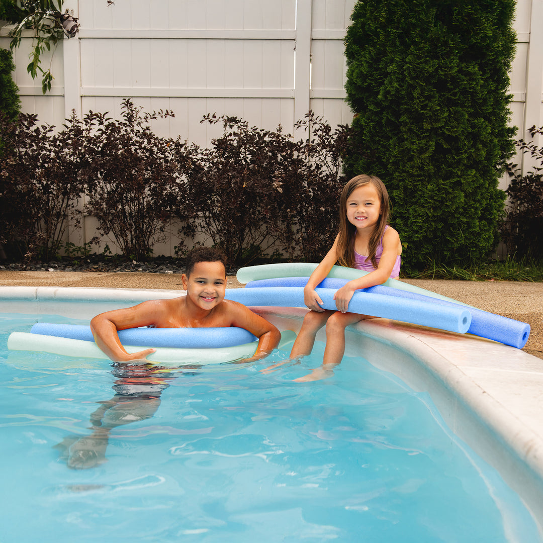 Two children sitting on a pool deck beside a swimming pool, using blue and white pool noodles. A white fence and green shrubs are in the background. 