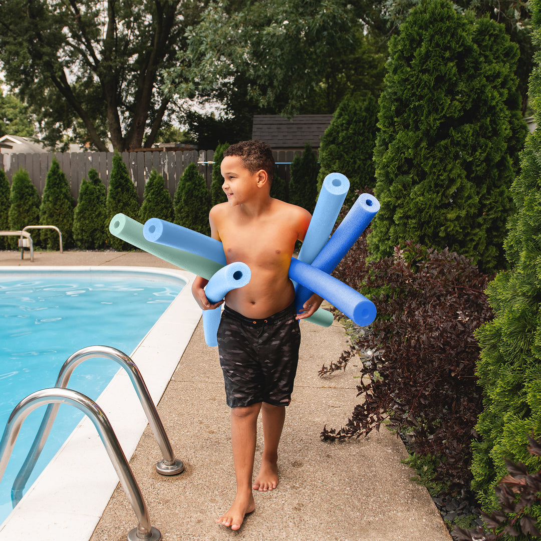 A young boy walking next to a swimming pool holding several blue and mint green pool noodles. 