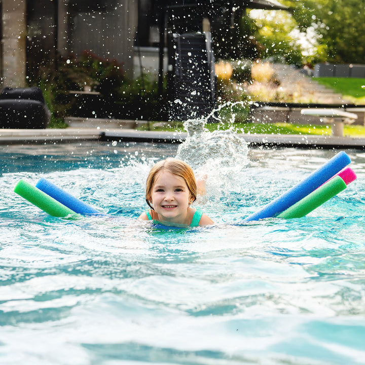 A young girl smiling in a swimming pool, using colorful foam pool noodles (blue, green, and pink) as flotation aids, with water splashing around her. 