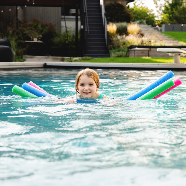 A young child wearing a blue swim ring float in a swimming pool, holding four colorful pool noodles (green, blue, pink, and blue) behind them. The background shows a backyard with a patio, stairs, and greenery. 