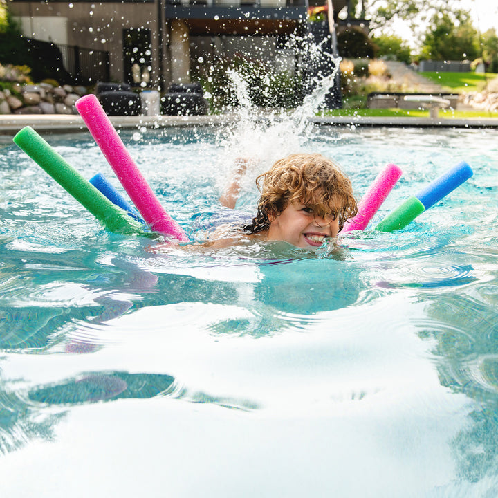 A child smiling and splashing in a swimming pool while using colorful foam pool noodles, including pink, green, blue, and a darker blue noodle. 