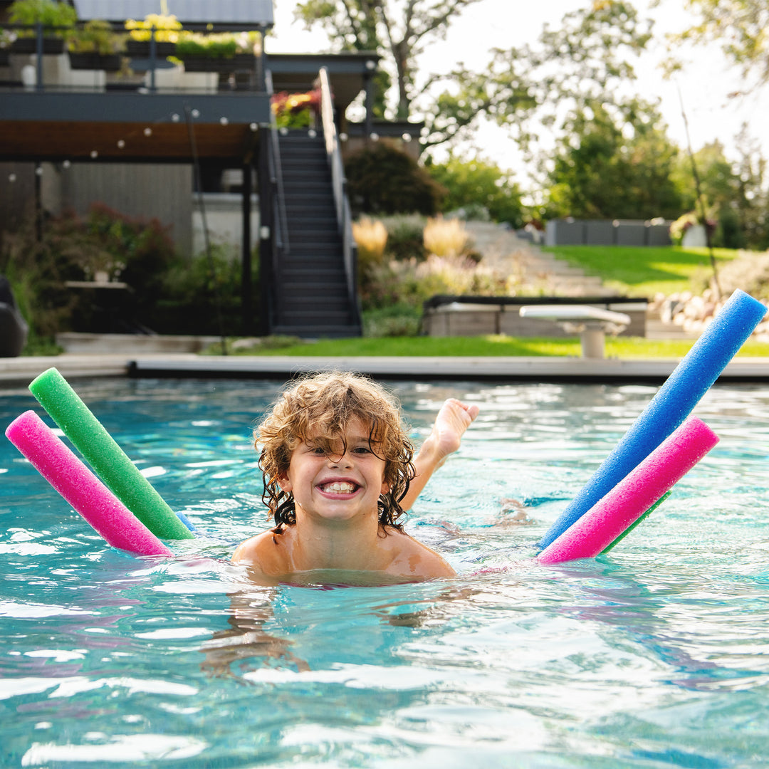 A smiling child with curly hair swims in a backyard pool, using four foam pool noodles in green, pink, blue, and pink. A modern house with a staircase and landscaped garden is visible in the background. 