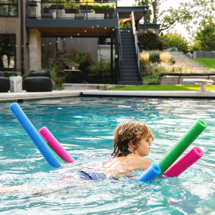 A child floating on a pool with four foam pool noodles in blue, pink, and green, with a modern house and outdoor patio in the background. 