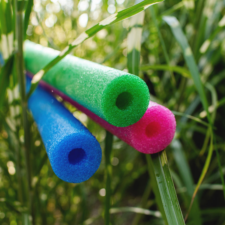 Three foam pool noodles in blue, green, and pink resting on tall green grass outdoors. 