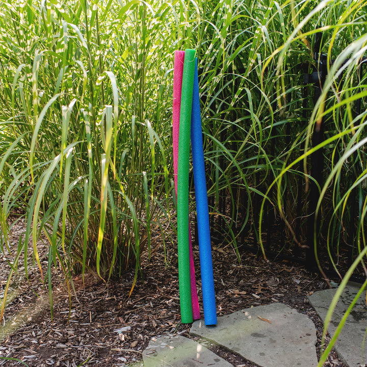 Three long, thin pool noodles in pink, green, and blue standing upright in a garden bed with tall green grass and mulch. 