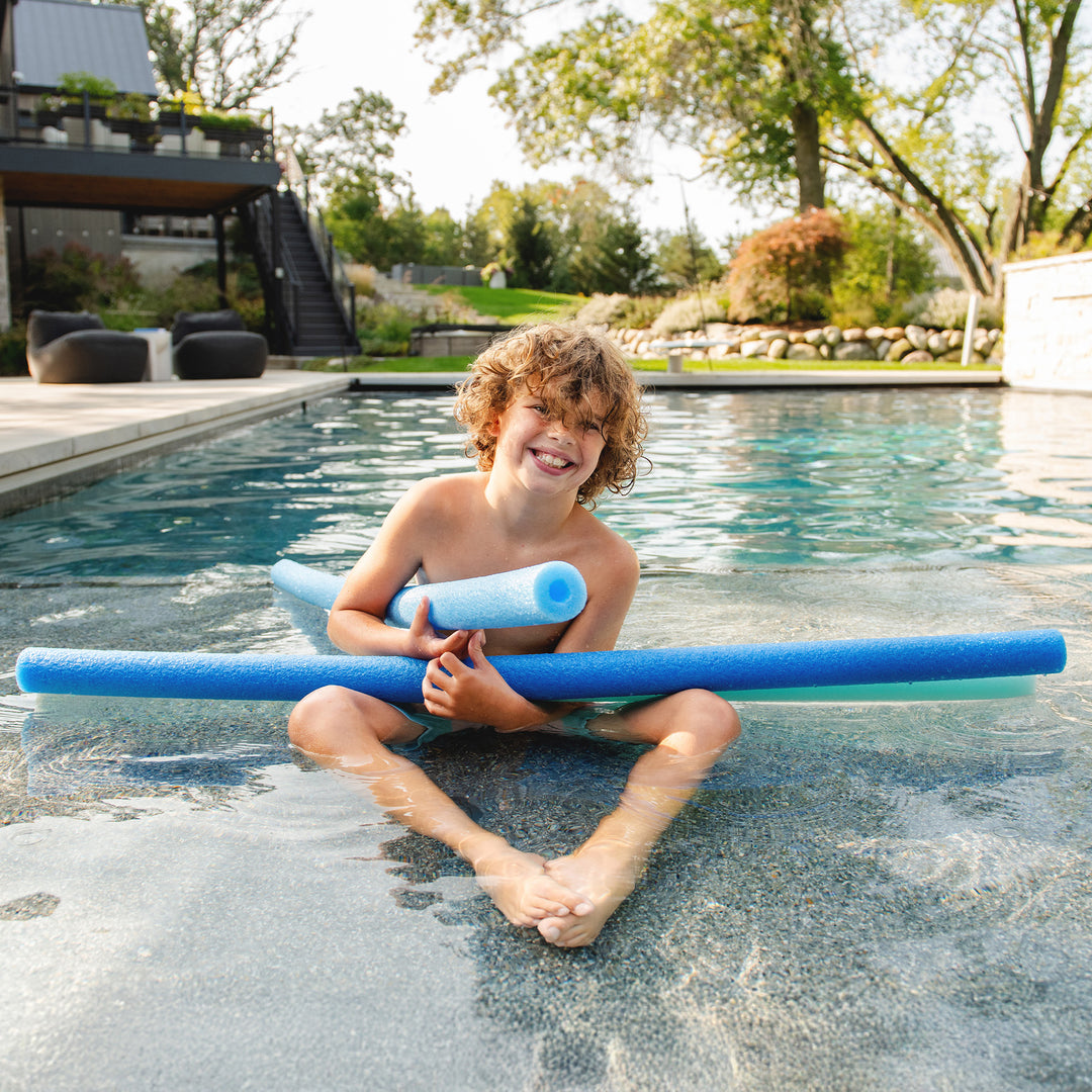 A smiling child sitting in a swimming pool holding a blue pool noodle, with another blue pool noodle resting behind them, in an outdoor backyard setting. 