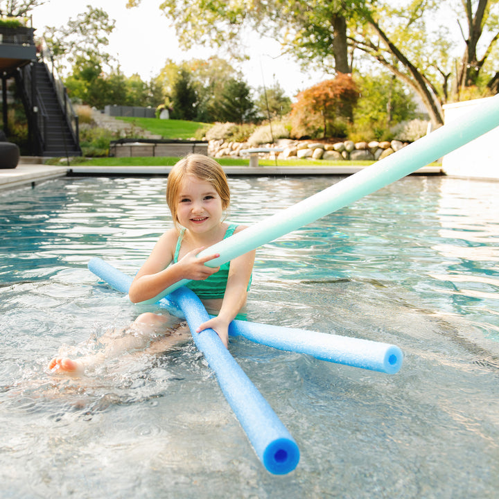 A young girl sitting in a swimming pool holding several blue and mint green pool noodles. 