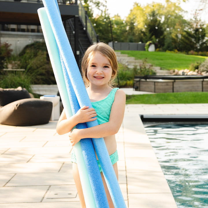 A young girl in a mint green swimsuit stands by a swimming pool holding three light blue pool noodles. 