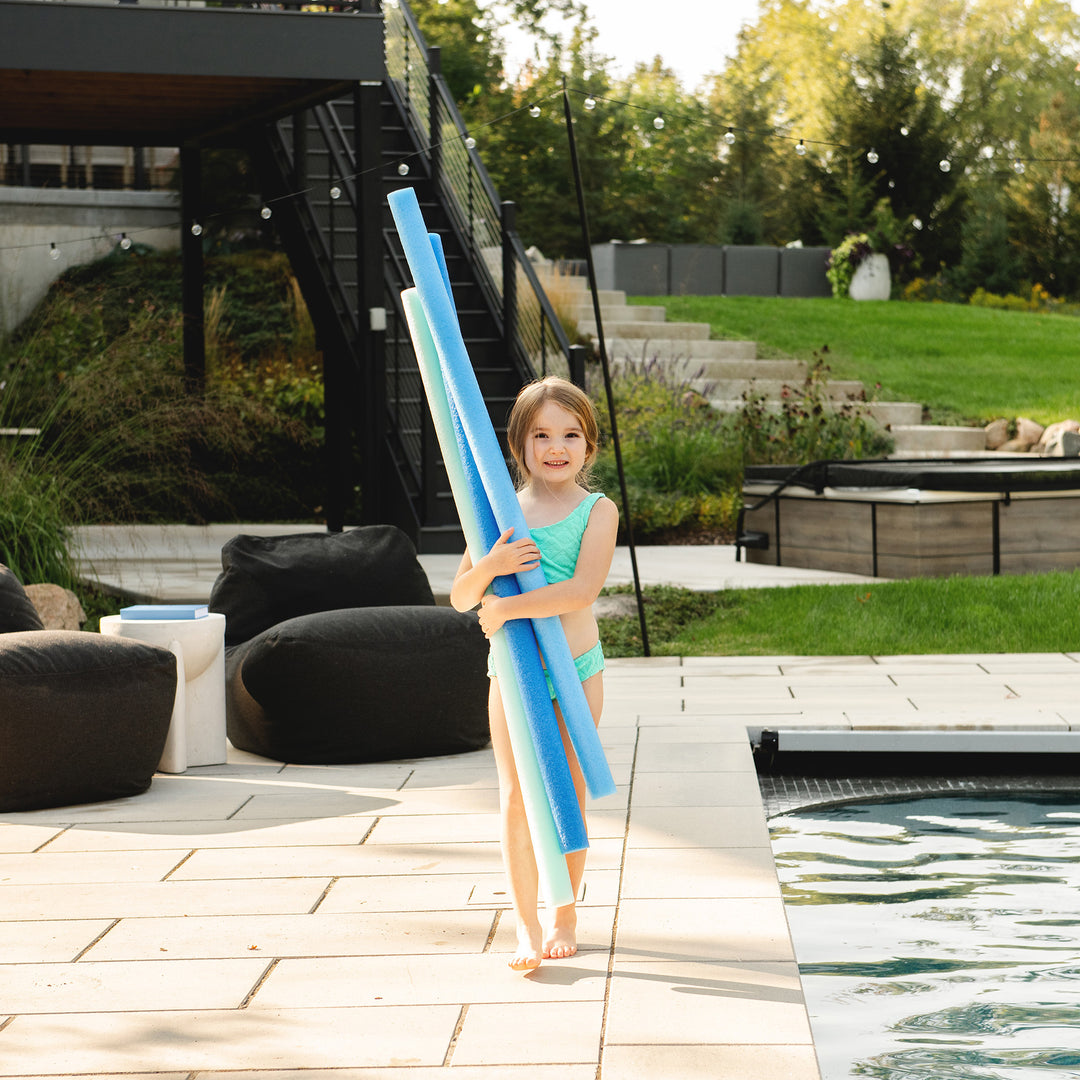 A young girl in a turquoise swimsuit stands on a poolside patio holding a stack of blue pool noodles. The background shows a modern outdoor setting with a staircase, lounge chairs, and a grassy lawn. 