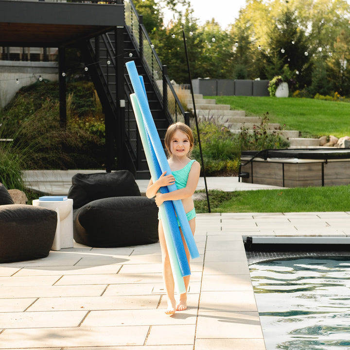 A young girl in a turquoise swimsuit stands on a poolside patio holding a stack of blue pool noodles. The background shows a modern outdoor setting with a staircase, lounge chairs, and a grassy lawn. 