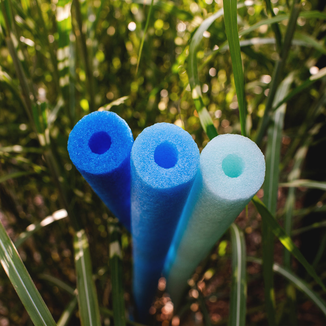 Three foam pool noodles in blue and light blue colors standing upright in tall green grass outdoors. 