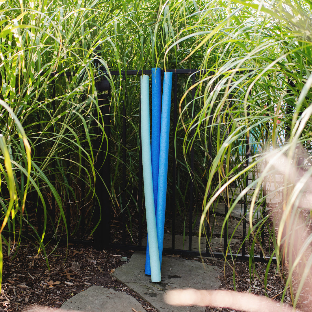 Three pool noodles in light blue, medium blue, and navy are standing vertically on a stone slab in a garden setting surrounded by tall green grass. 