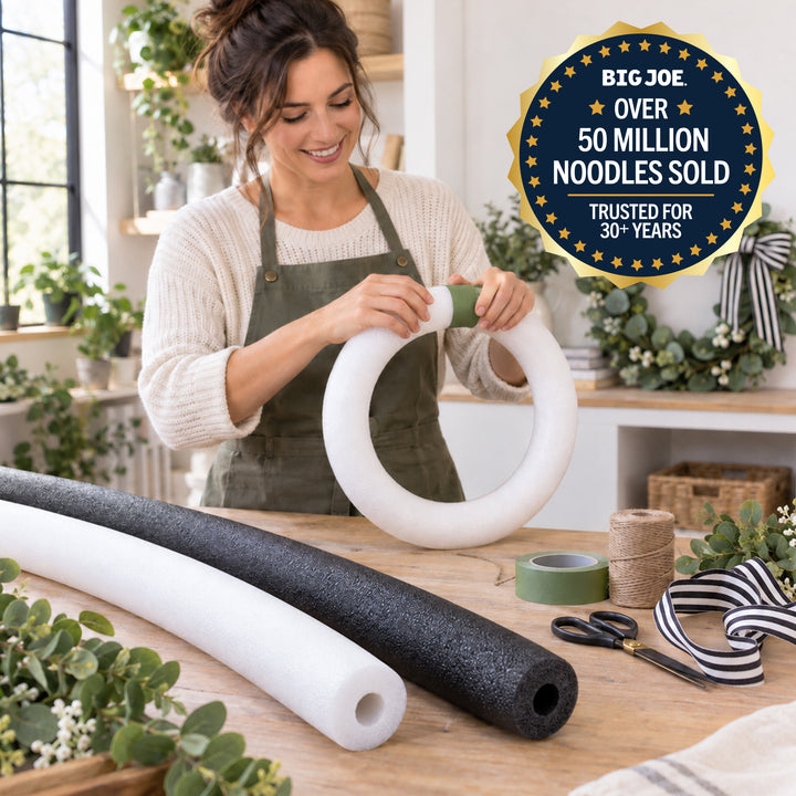 A woman in an apron is assembling a foam flower head on a wooden table in a bright florist shop. Two foam noodles, one white and one black, are laid out in the foreground, along with green tape, twine, scissors, and decorative plants. 