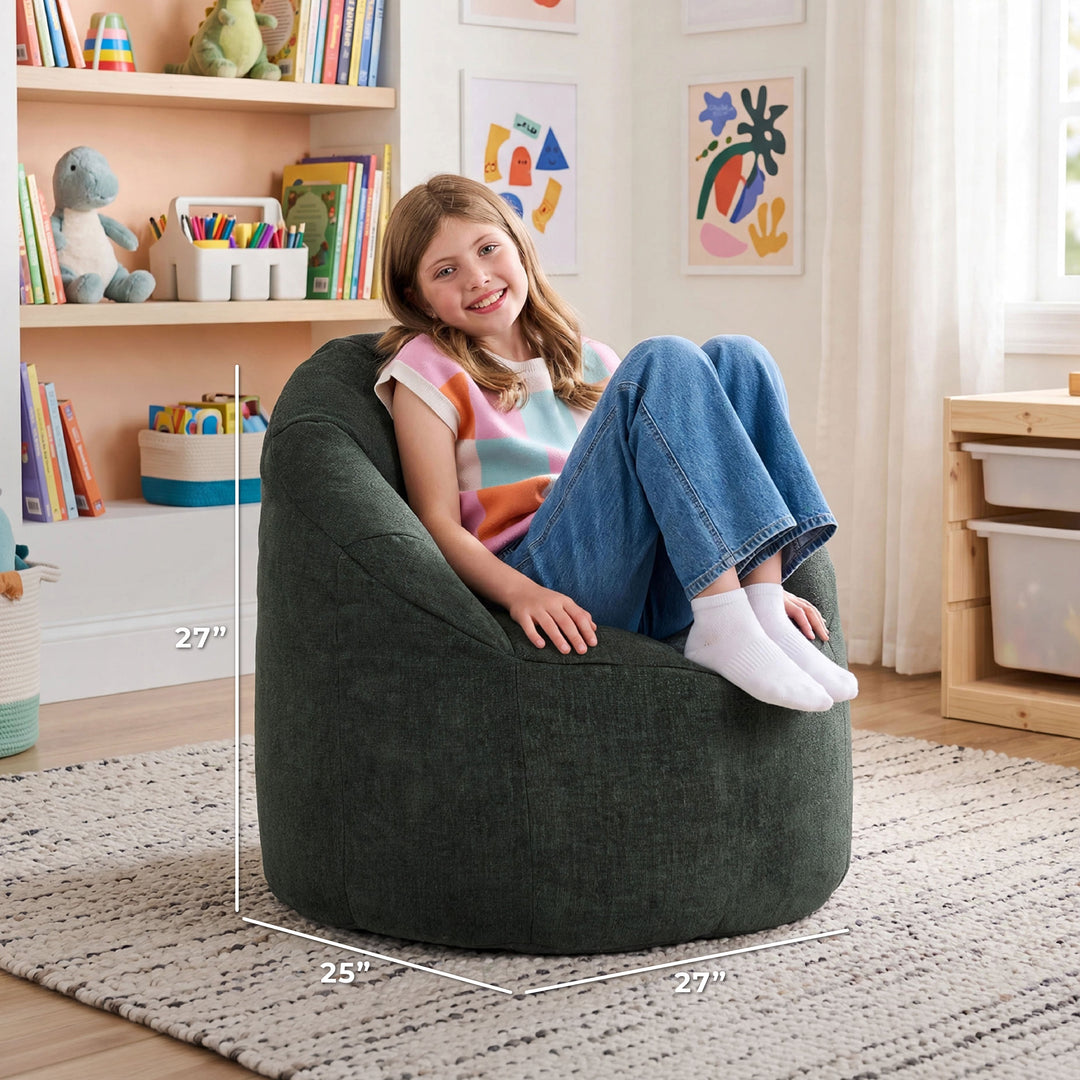A child sits in a large dark green bean bag chair in a bright, styled room. White dimension lines overlay the chair showing its measurements: 27 inches high, 25 inches wide, and 27 inches deep. 