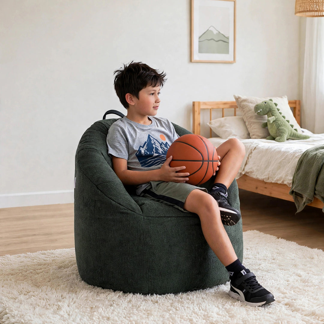 A boy sits on a dark green bean bag chair in a bright bedroom, holding an orange basketball. The bean bag chair is upholstered in a textured dark green fabric and is placed on a white shag rug. 