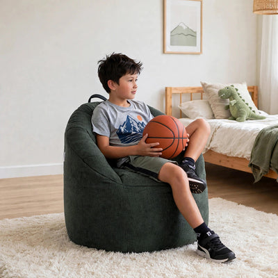 A boy sits on a dark green bean bag chair in a bright bedroom, holding an orange basketball. The bean bag chair is upholstered in a textured dark green fabric and is placed on a white shag rug. 