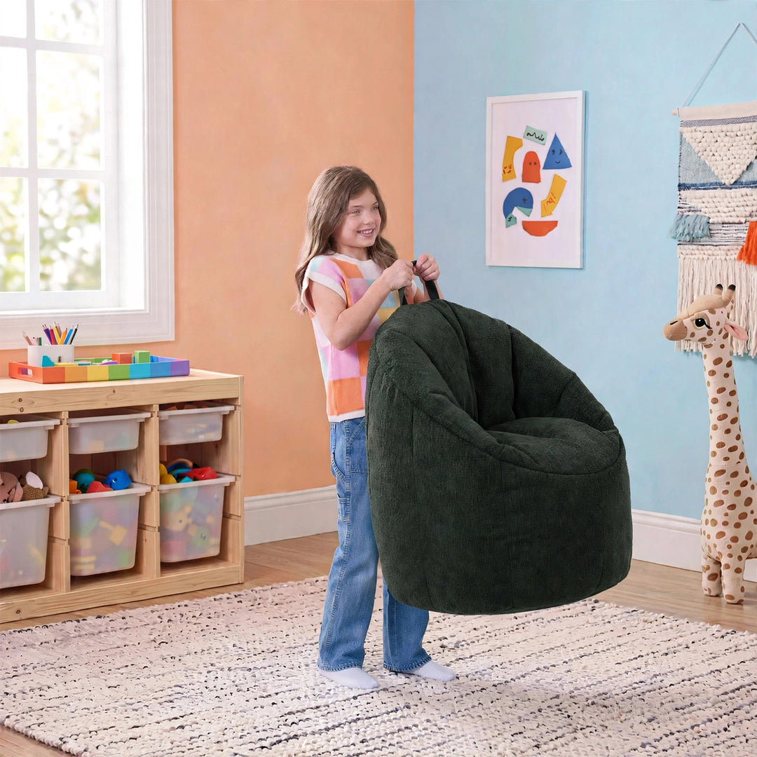 A young girl stands in a brightly lit playroom holding a large, plush dark green bean bag armchair. The room features peach and light blue walls, a wooden toy organizer, and a giraffe toy. 
