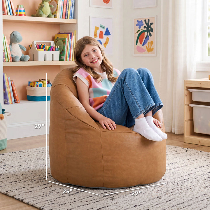 A child sits on a large tan bean bag chair in a bright playroom. White dimension lines on the image show the chair&