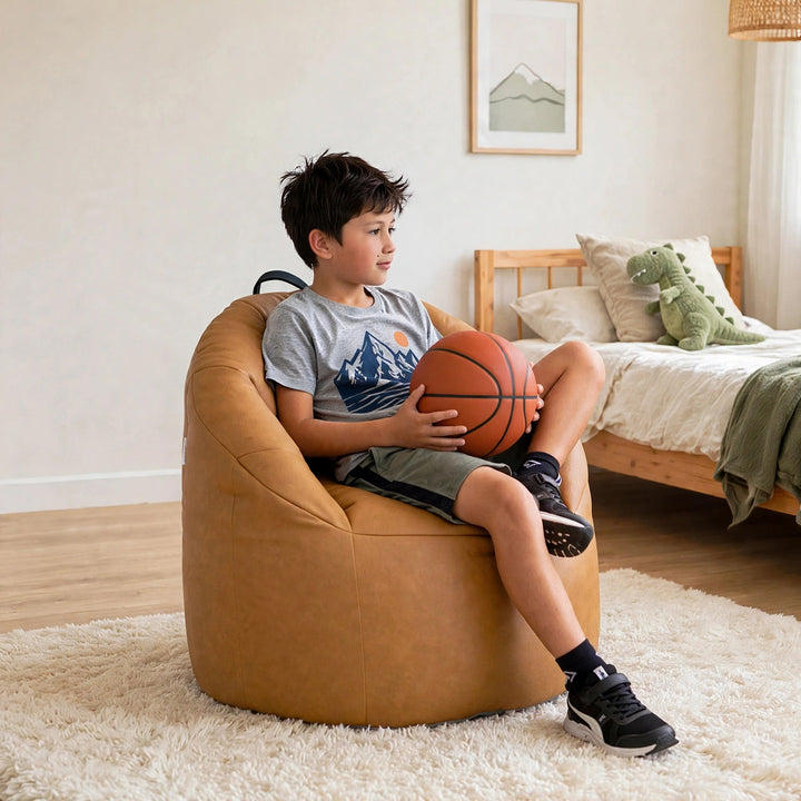 A child sits in a tan oversized bean bag chair in a bright bedroom, holding an orange basketball. The room has light wood flooring, a white shag rug, a wooden bed, and a green dinosaur plush toy. 