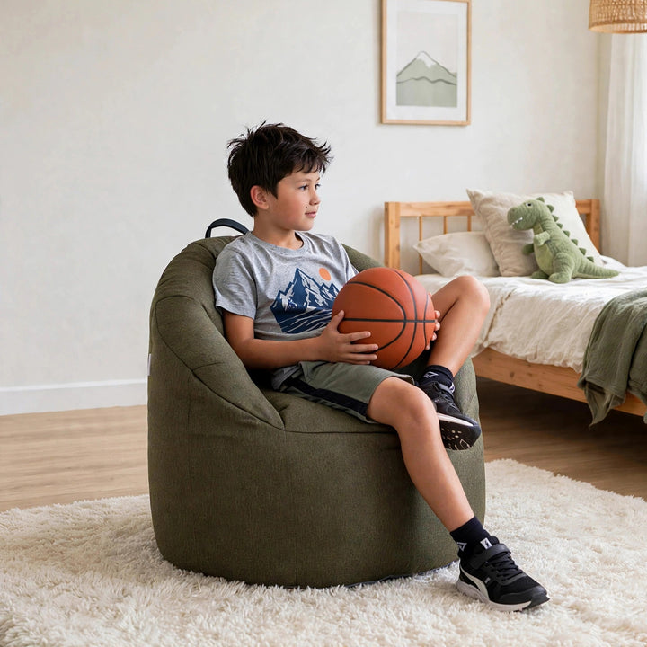 A boy sits in a large olive green bean bag chair, holding an orange basketball in a bright, modern bedroom. 