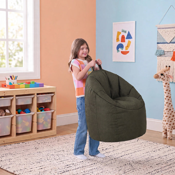 A young girl stands in a playroom holding a large, dark olive green bean bag chair. The room features a peach-colored wall on the left, a light blue wall on the right, a wooden toy organizer, and a giraffe plush toy. 