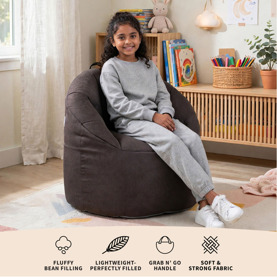 A young girl wearing a gray sweatshirt and matching sweatpants sits smiling in a large, dark brown bean bag chair in a bright, modern children&