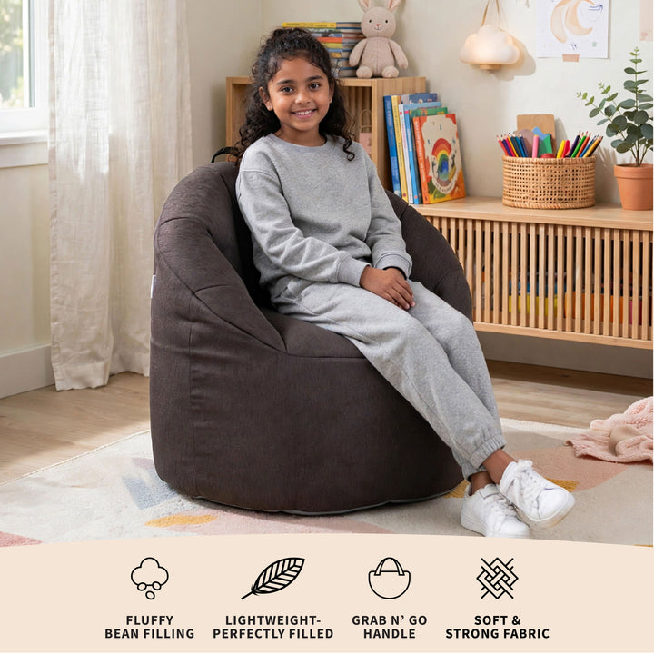 A young girl wearing a gray sweatshirt and matching sweatpants sits smiling in a large, dark brown bean bag chair in a bright, modern children&