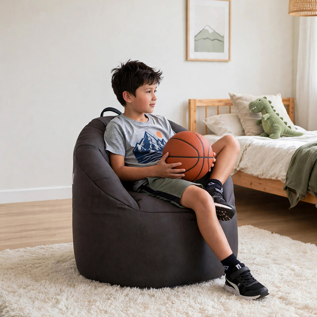 A boy sits in a dark gray oversized bean bag chair in a bright bedroom, holding an orange basketball. The room has light wood flooring, a white shag rug, a wooden bed, and a green dinosaur plush toy. 