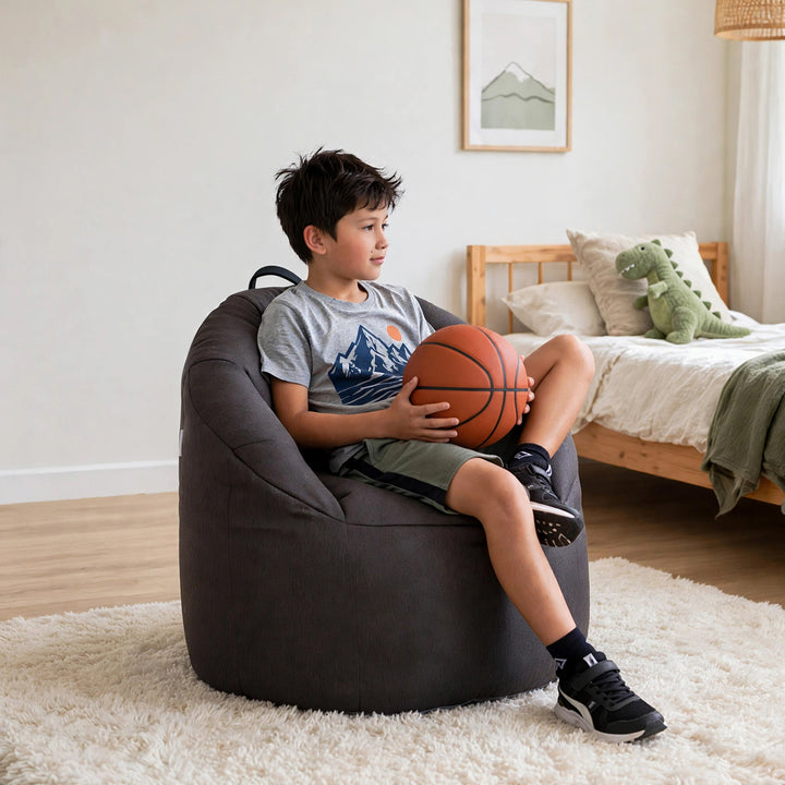A boy sits in a dark gray oversized bean bag chair in a bright bedroom, holding an orange basketball. The room has light wood flooring, a white shag rug, a wooden bed, and a green dinosaur plush toy. 