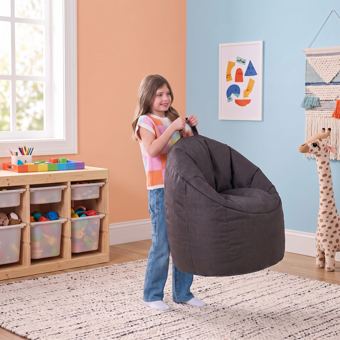 A young girl stands in a brightly lit playroom holding a large, dark gray fabric bean bag chair. The room features a peach-colored wall on the left, a light blue wall on the right with a colorful framed art print, and a wooden toy organizer cabinet on the left. 