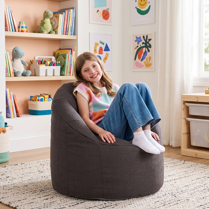 A young girl with long brown hair sits smiling in a large, round, dark brown bean bag chair in a brightly lit children&