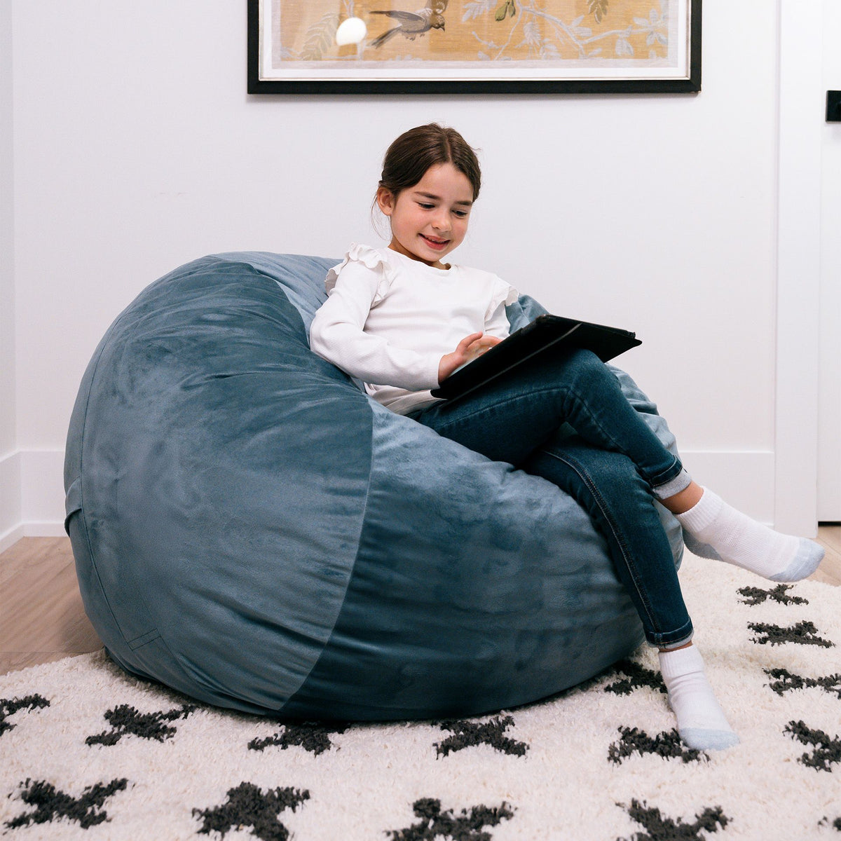 Child sitting on a large blue bean bag reading a book in a cozy room. 